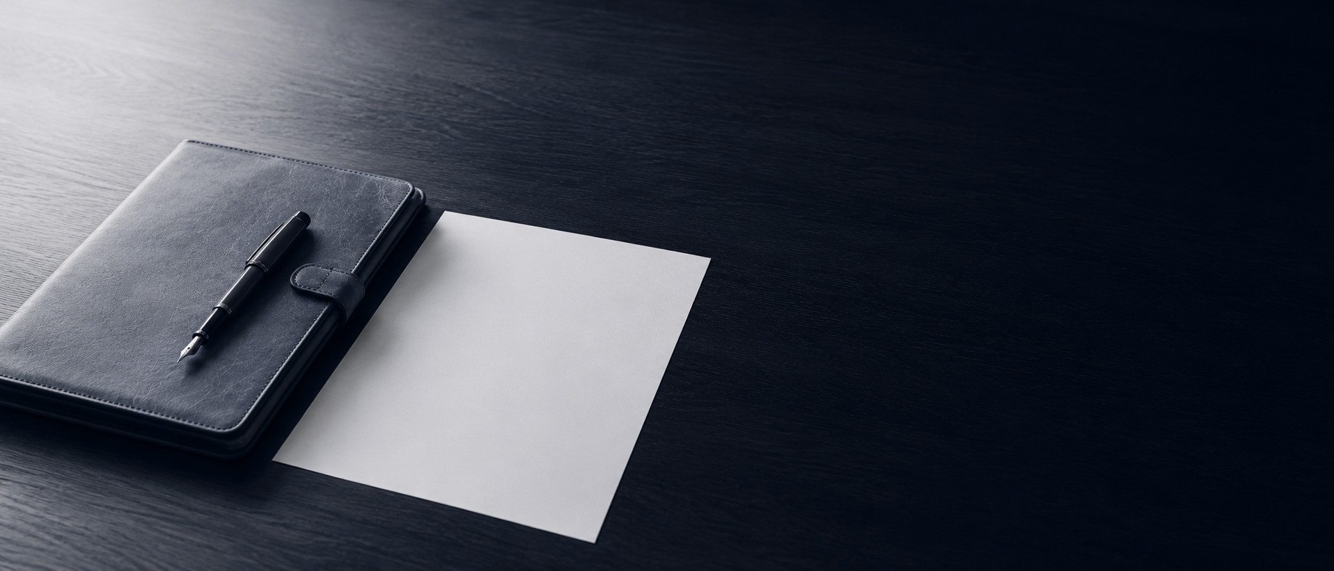A leather journal, fountain pen, and a blank sheet of paper on a dark desk